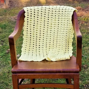 Pu&ograve; includere: Una coperta all'uncinetto di colore giallo pallido con un bordo a festoni &egrave; appoggiata su una sedia a dondolo in legno. La sedia &egrave; davanti a uno sfondo erboso.