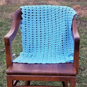 Pu&ograve; includere: Una coperta all'uncinetto di colore azzurro chiaro &egrave; appoggiata su una sedia a dondolo in legno. La coperta ha un motivo semplice e ripetitivo.