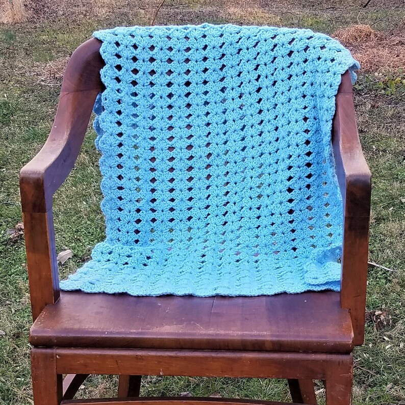 Pu&ograve; includere: Una coperta all'uncinetto di colore azzurro chiaro &egrave; appoggiata su una sedia a dondolo in legno. La coperta ha un motivo semplice e ripetitivo.