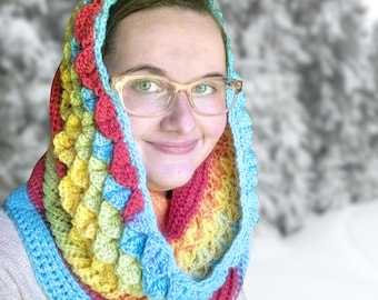 May include: A colorful, crocheted cowl in a rainbow of hues, including red, yellow, blue, and green. The cowl features a textured, scale-like pattern and a scalloped edge. The handmade accessory is designed to be worn around the neck and head.