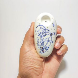 May include: A smooth, off-white ceramic pipe featuring a hand-painted blue line drawing of a cat wearing a space helmet and suit. The design includes small gold-colored dots and blue star shapes scattered around the cat on the rounded surface.