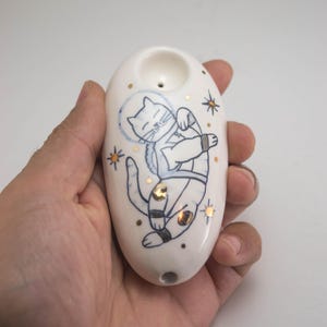 Könnte beinhalten: Eine ovale, weiße Keramikpfeife mit einer handgemalten blauen Strichzeichnung einer Katze in einem Weltraumhelm. Das Design enthält kleine goldfarbene Akzente auf dem Körper der Katze und umgebende Sterne auf der glänzenden weißen Oberfläche.