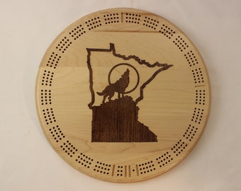 May include: A circular wooden cribbage board featuring a design of the state of Minnesota with a wolf howling at the moon. The board has a dark brown stain and is surrounded by a ring of holes for pegs.