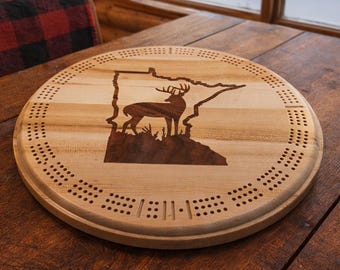May include: A circular wooden cribbage board featuring a laser-cut outline of Minnesota and a deer silhouette. The board is dotted with holes for pegs and rests on a wooden table. The background shows a window with a snowy scene.