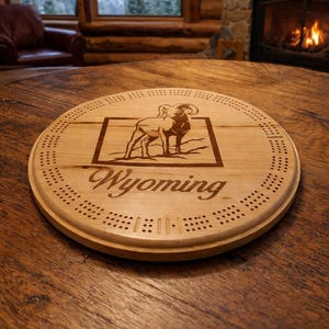May include: A round wooden cribbage board with a laser-etched image of a ram inside a rectangular frame. The word "Wyoming" is etched below the image. The board has numerous holes for pegs and is set on a wooden table.