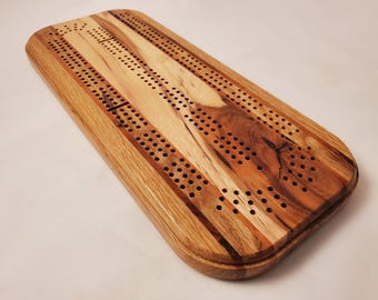 May include: A wooden cribbage board with a rectangular shape and rounded corners. The board features a natural wood grain pattern with various shades of brown and tan. Numerous holes are drilled for peg placement, and the board is designed for the classic card game.