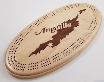 Puede incluir: Tablero de cribbage ovalado de madera con un mapa de Anguila en madera oscura sobre un fondo claro. La palabra "Anguilla" está escrita. El tablero tiene muchos agujeros para clavar y puntuar.