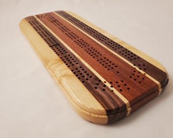 May include: Wooden cribbage board with a rectangular shape and rounded edges. The board features three parallel stripes of light, medium, and dark wood, with drilled holes for pegs. A classic game for two or more players.