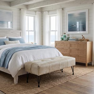 May include: A bedroom featuring a white wooden bed frame with blue and white bedding, a light-colored tufted bench with black metal legs, and a six-drawer wooden dresser with woven rattan panels, all set on a textured natural fiber area rug.