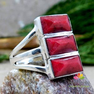 Könnte beinhalten: Ein Silberring mit drei rechteckigen, facettierten roten Edelsteinen. Der Ring hat ein geteiltes Schaftdesign und ist auf einem strukturierten Stein platziert. Die Edelsteine haben eine leuchtend rote Farbe.