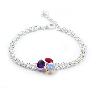 May include: A silver bracelet with three colorful gemstones: a teardrop-shaped purple amethyst, a round red ruby, and an oval opal. The bracelet features a double chain design and a lobster clasp.