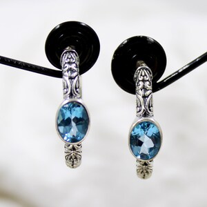 May include: A pair of silver hoop earrings with a blue topaz gemstone in each earring. The earrings have a detailed filigree design.