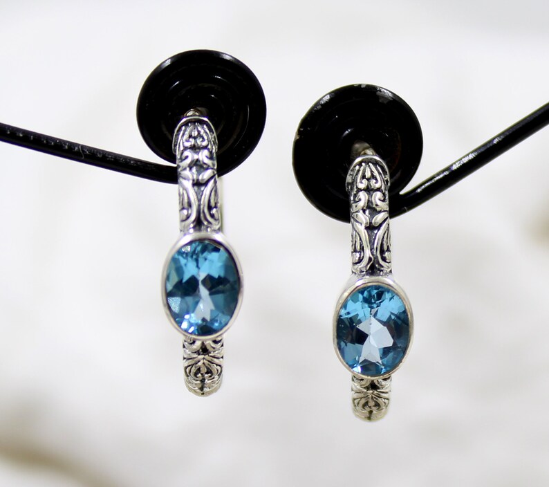 May include: A pair of silver hoop earrings with a blue topaz gemstone in each earring. The earrings have a detailed filigree design.