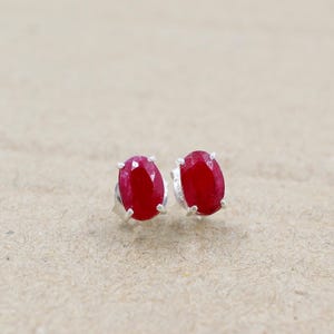 May include: A pair of oval-shaped ruby stud earrings. The rubies are a deep red color and set in a silver metal. The earrings are displayed on a neutral background.