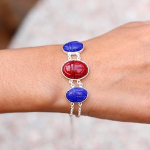 May include: A silver bracelet featuring three oval-shaped scarab beetle charms. The central charm is red, while the top and bottom charms are blue. The bracelet is worn on a wrist.