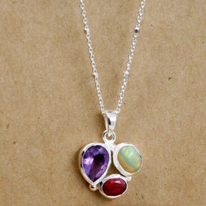 Könnte beinhalten: Eine silberne Anhängerkette mit drei Edelsteinen: einem birnenförmigen, lila Amethyst, einem ovalen, roten Rubin und einem runden Opal mit grün-gelben Farbtönen. Die Halskette hat eine zarte Silberkette.