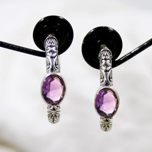 May include: A pair of silver hoop earrings with a detailed floral design and a light purple amethyst gemstone in each hoop.