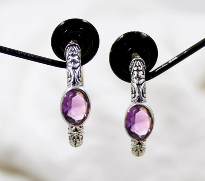 May include: A pair of silver hoop earrings with a detailed floral design and a light purple amethyst gemstone in each hoop.