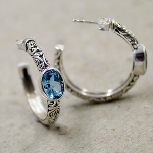 May include: A pair of silver hoop earrings with a blue topaz gemstone set in each earring. The earrings have an intricate floral design.