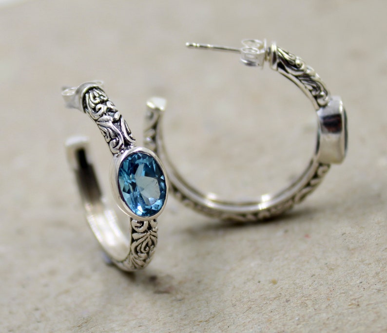 May include: A pair of silver hoop earrings with a blue topaz gemstone set in each earring. The earrings have an intricate floral design.