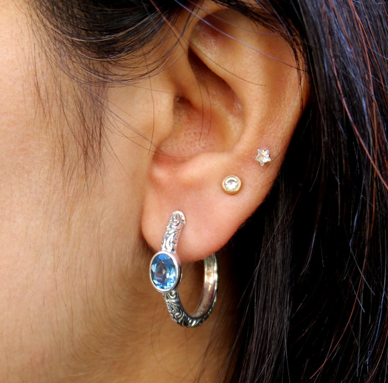 May include: Silver hoop earrings with a blue gemstone and intricate floral designs. The earrings are worn in a person's ear.