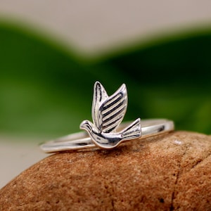 May include: A silver ring featuring a dove design. The dove is in flight with its wings outstretched. The ring is resting on a brown rock.