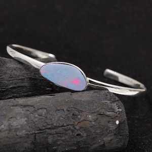 May include: A silver bangle bracelet featuring a single oval-shaped opal gemstone set in the centre. The opal displays a vibrant blue-green colour with flashes of red and pink.
