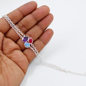 May include: A silver bracelet featuring three colorful gemstones: a purple amethyst, a red ruby, and a blue opal. The bracelet has a delicate chain design and is displayed against a white background. This piece of jewelry is suitable for everyday wear or special occasions.