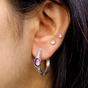 May include: Silver hoop earrings with an amethyst gemstone. The earrings have an intricate floral design.