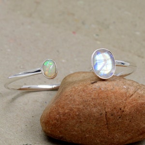 May include: A silver cuff bracelet with two gemstones. One is an oval opal with iridescent flashes, and the other is a moonstone with a blue and white sheen. The bracelet is displayed on a brown rock.