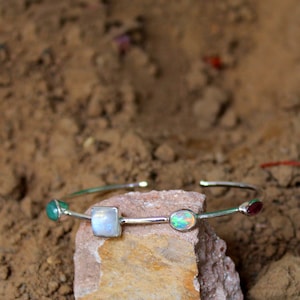 May include: A silver bangle bracelet featuring four gemstones: a green stone, a white moonstone, a white opal, and a red stone. The bracelet is resting on a rough, brown rock.