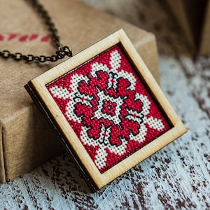 May include: A red and white embroidered design in a wooden frame, hanging from a bronze chain. The design is a traditional folk pattern with a floral motif.