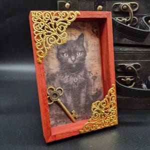 May include: A rectangular red wooden frame featuring ornate gold-toned metal filigree corner accents. Inside the frame is a sepia-toned print of a black cat wearing a collar, with a vintage-style metal skeleton key attached to the surface.