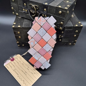 May include: A decorative mosaic art piece in shades of pink, orange, and gray, with a woven pattern. The art piece is displayed in front of two dark wooden treasure chests with brass accents. A tag is attached to the art piece.