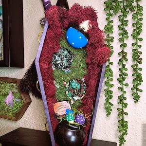May include: A coffin-shaped decorative piece with a purple frame, filled with moss, decorative coins, and a small cauldron with mushrooms. A blue egg with white polka dots and a witch's broom are also visible. The theme is mystical and witchy.