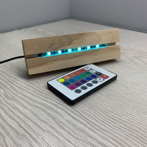 Könnte beinhalten: Ein Holzsockel mit einem LED-Lichtstreifen, der mit einer Fernbedienung gesteuert werden kann. Die Fernbedienung hat Tasten für verschiedene Farben, Modi und Helligkeiten.