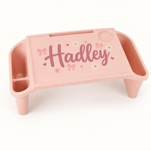 May include: A light pink plastic lap desk featuring the name Hadley printed in a dark pink script font. The surface includes a slot for a tablet or phone, a circular cup holder, and decorative graphics of small white daisies and pink bows.