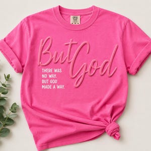 May include: A bright pink short-sleeved t-shirt featuring the raised, embossed text 'But God' in a cursive font. Below this, the text 'THERE WAS NO WAY. BUT GOD MADE A WAY.' is printed in a clean, white sans-serif font on the front of the garment.