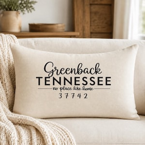 May include: A rectangular off-white throw pillow featuring black text that reads Greenback TENNESSEE no place like home 37742. The pillow has a smooth, woven fabric texture and is positioned on a light-colored sofa with a textured throw blanket.