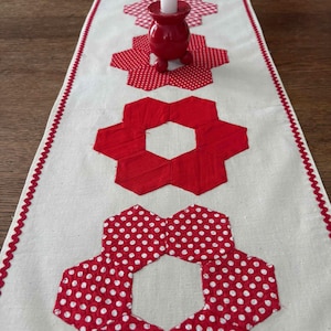 May include: A long, off-white fabric table runner featuring a repeating pattern of red and white polka-dot and solid red hexagonal flower motifs. The runner has a red zigzag stitched border and holds a red candle holder with a white candle.