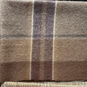 May include: Close-up of a folded, textured fabric with a plaid pattern. The fabric features shades of brown, tan, and a deep burgundy. Vertical stripes of white and burgundy intersect the plaid design. The fabric appears to be wool or a similar material.