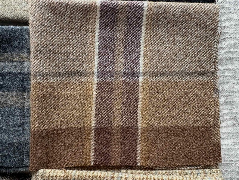 May include: Close-up of a folded, textured fabric with a plaid pattern. The fabric features shades of brown, tan, and a deep burgundy. Vertical stripes of white and burgundy intersect the plaid design. The fabric appears to be wool or a similar material.
