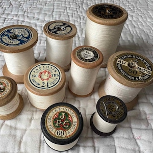 Peut inclure: Plusieurs bobines de fil en bois anciennes, remplies de fil blanc. Les bobines présentent des étiquettes imprimées avec du texte et des motifs, dont "J&P Coats" et des mesures en yards. Les bobines sont disposées sur un tissu blanc texturé.
