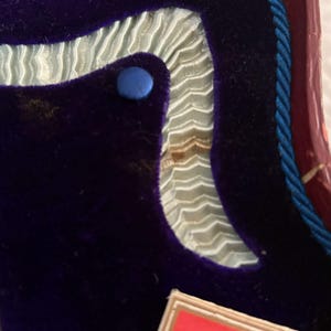May include: Close-up of a section of a box interior. The interior features a deep purple velvet material, a light blue ribbon trim, and a blue button. A red and blue rope trim is visible.