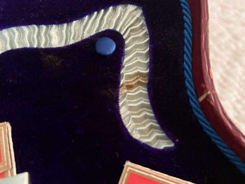 May include: Close-up of a section of a box interior. The interior features a deep purple velvet material, a light blue ribbon trim, and a blue button. A red and blue rope trim is visible.