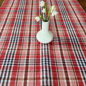 May include: A red, white, and navy blue plaid table runner with a white vase holding white flowers. The vase has a textured surface and is centered on the runner. The runner is made of woven fabric.