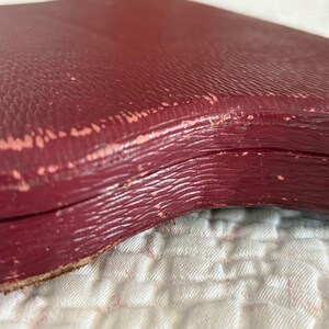 May include: Close-up of a burgundy-colored, vintage box with a textured surface. The box appears to be made of leather or a similar material, with visible wear and tear along the edges, revealing a lighter pinkish layer.