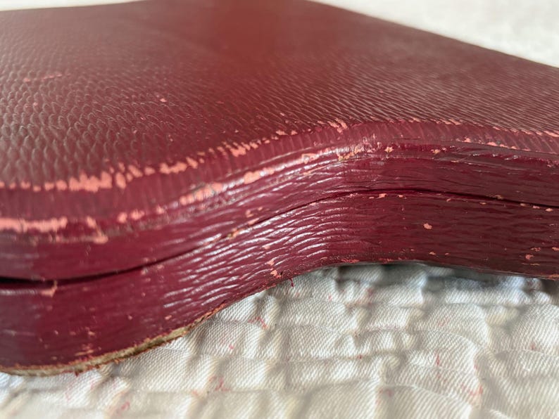 May include: Close-up of a burgundy-colored, vintage box with a textured surface. The box appears to be made of leather or a similar material, with visible wear and tear along the edges, revealing a lighter pinkish layer.