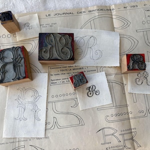 May include: A collection of vintage woodblock printing stamps featuring the letter "R" in various styles. The stamps are arranged on a white surface with a sheet of paper showing a drawn outline of the letter "R". The paper has the text "LE JOURNAL DES BRODEUSES - N 783 - 1 Juin 1960" printed on it.
