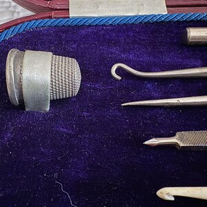 May include: A vintage sewing kit with a silver thimble, needles, and crochet hooks, set against a deep purple velvet background. The tools are neatly arranged within a red and blue-trimmed case, showcasing craftsmanship.